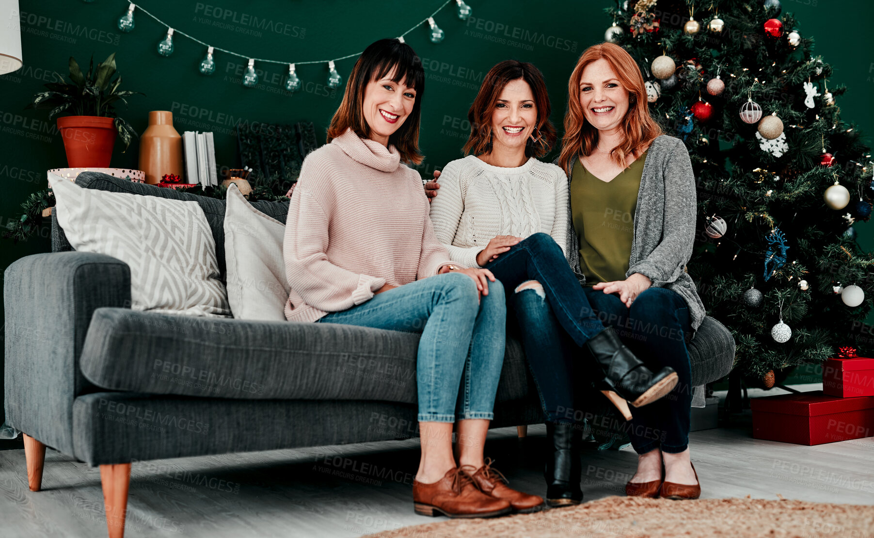 Buy stock photo Portrait of three attractive middle aged women seated together on a sofa with a Christmas tree in the background at home