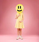 Emoji face, balloon and child in studio, covering and hiding against a pink background space. Happy, smile and girl holding toy, game and playing while posing behind inflatable, fun and innocent