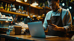 Laptop, phone and barista in coffee shop working at night on stock inventory for startup. Technology, small business and man waiter with cellphone and computer for online website in cafe restaurant.