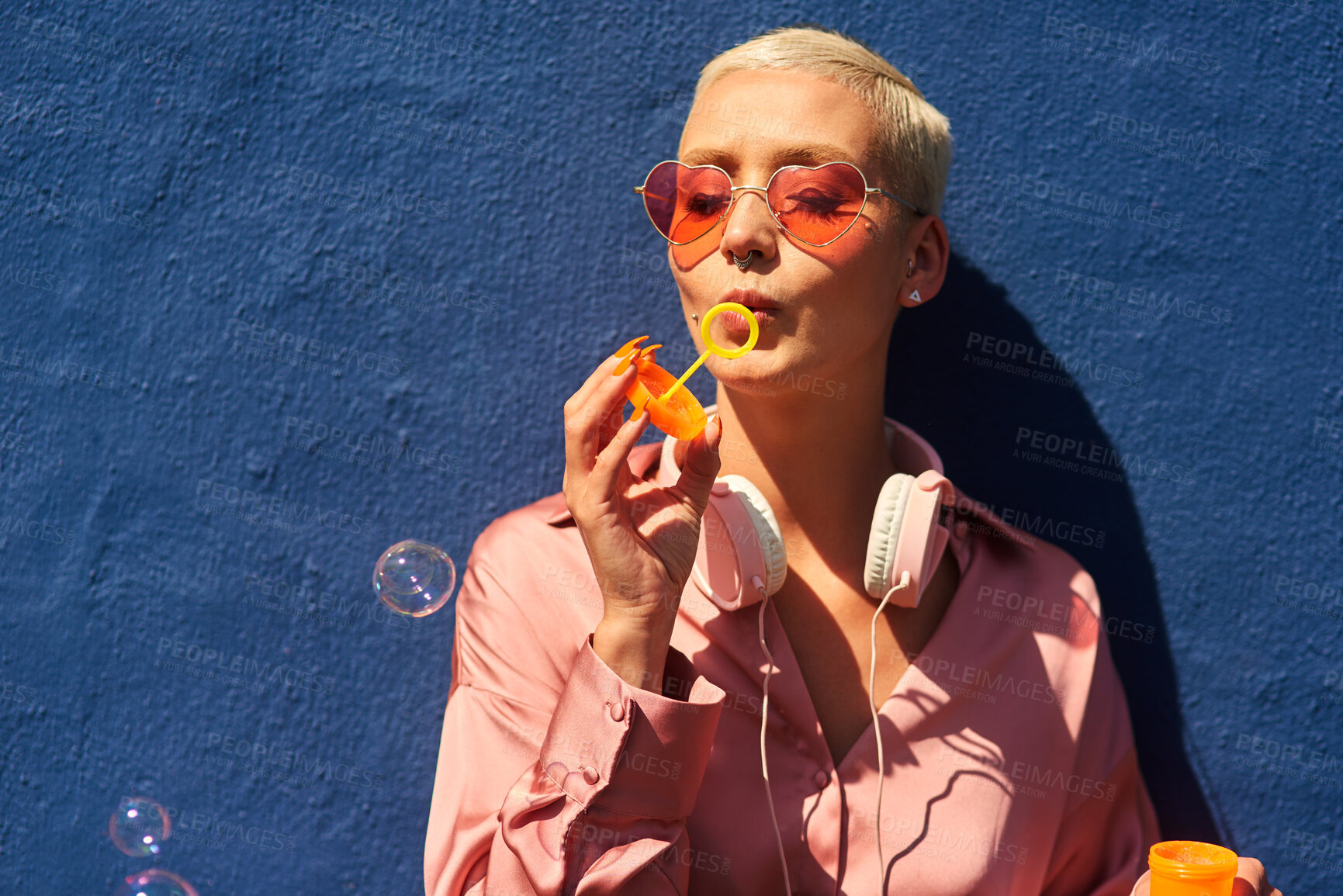 Buy stock photo Young, woman and blowing bubbles against wall for fashion or wellness, cool with heart sunglasses for trend. Female person, outdoor and backdrop with sunshine, headphones and clothes for style in USA