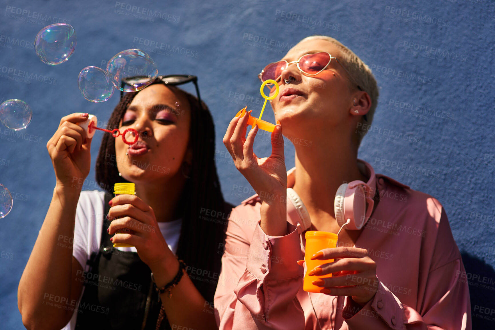 Buy stock photo Women, blowing bubbles and fun at wall with urban fashion, streetwear and nostalgia in city. Girl friends, gen z style and relax in sunshine with game, playful and sunglasses with toy for bonding