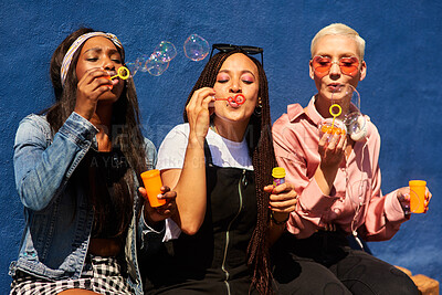 Buy stock photo Blowing bubbles, women and fun at wall with urban fashion, streetwear and nostalgia in city. Girl friends, gen z style and relax in sunshine with game, playful and sunglasses with toy for bonding