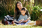 A little girl and her mother reading a book while relaxing in a park or garden. Young woman with a little child learning and getting an education, sitting on the grass in nature and having fun