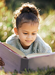 A little girl reading a book while relaxing in a park or garden. Mixed race child learning and getting an education, lying on the grass in nature and having fun