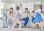 Stressed parents sitting on couch with children running around.Excited children playing around upset parents. Frustrated couple children running around. Unhappy parents upset at kids. Children playing