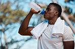 Young active black male athlete holding a bottle and drinking water with a towel around his neck in a forest outside. Runny feeling dehydrated quenching his thirst with some water on sunny hot summers day