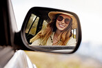 Reflection in the rear view mirror of a young beautiful woman's car while travelling to a vacation. Caucasian woman wearing sunglasses and a hat inside a car going on a roadtrip looking at her reflection and smiling while driving on the road