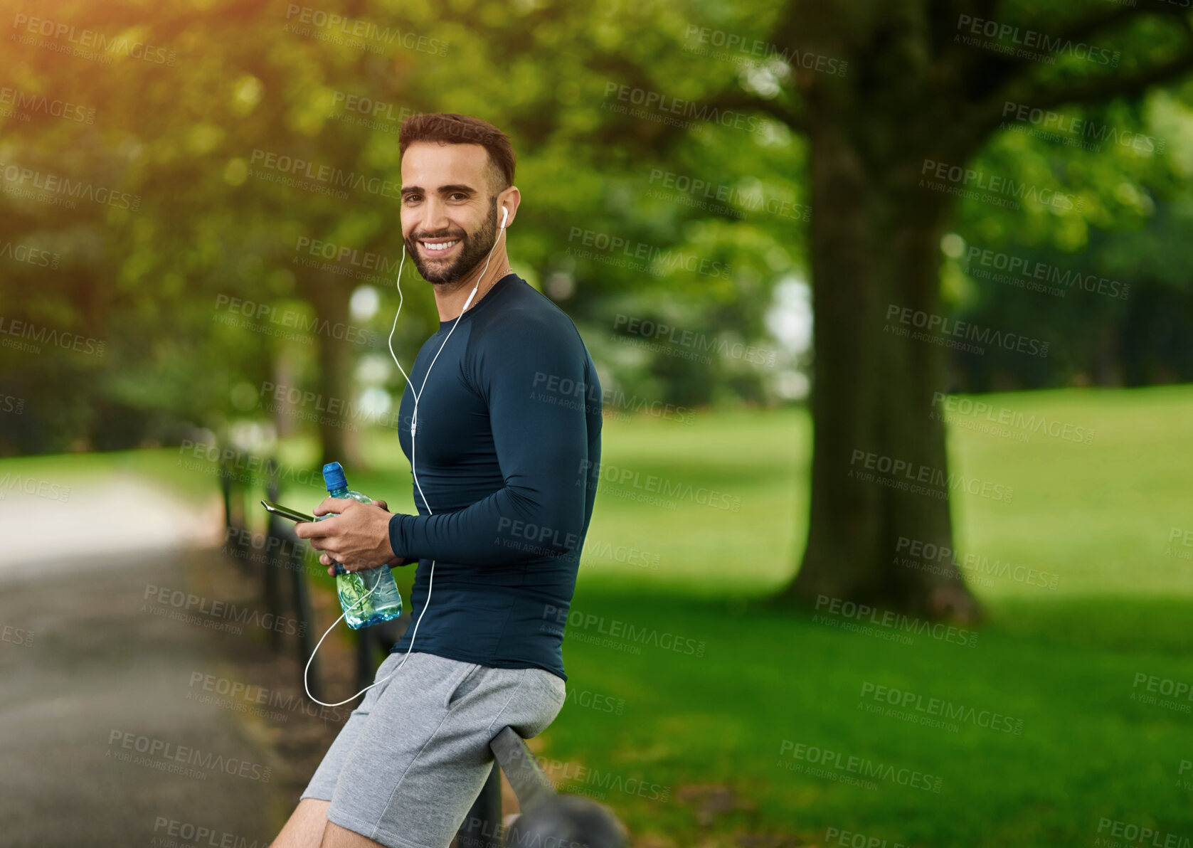 Buy stock photo Man, park and runner with earphones in portrait with smile, water or phone for audio streaming app. Person, happy and smartphone with bottle, fence and online subscription to listen to music on break