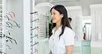 Woman, glasses and choice by shelf with optometry, thinking and smile for trendy style with vision. Person, customer experience and decision for eye care, health and wellness with retail sale in shop