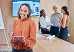 Young mixed race businesswoman with curly hair holding and working on a digital tablet while standing in a meeting at work. One hispanic female boss smiling while using a digital tablet in a boardroom