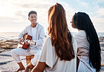 A young man at the beach playing his guitar with his two female friends. Diverse group of friends listening music on a sunny day