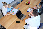 Family working at a table from above, doing homework at home. A young man using a laptop and woman helping. A little boy and girl playing while their father is online. Remote learning in quarantine