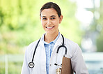 Confident young mixed race female doctor standing with clipboard a medical office. One hispanic woman in a white coat with stethoscope. Trusted practitioner caring for the health of patients