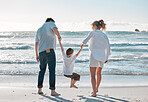 Caucasian family from behind sharing quality time while enjoying a fun summer vacation at the beach. Loving parents swinging their playful little son by the arms while bonding and walking on the shore