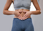 Closeup of one fit caucasian woman framing hands around her slim belly to show weight loss while exercising against a grey background. Female athlete with toned body caring for gut digestion and wellbeing through active lifestyle and healthy diet