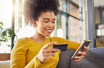 Young happy mixed race woman using a credit card and phone alone at home. Content hispanic female with a curly afro making an online purchase with a debit card and cellphone while sitting on the couch at home