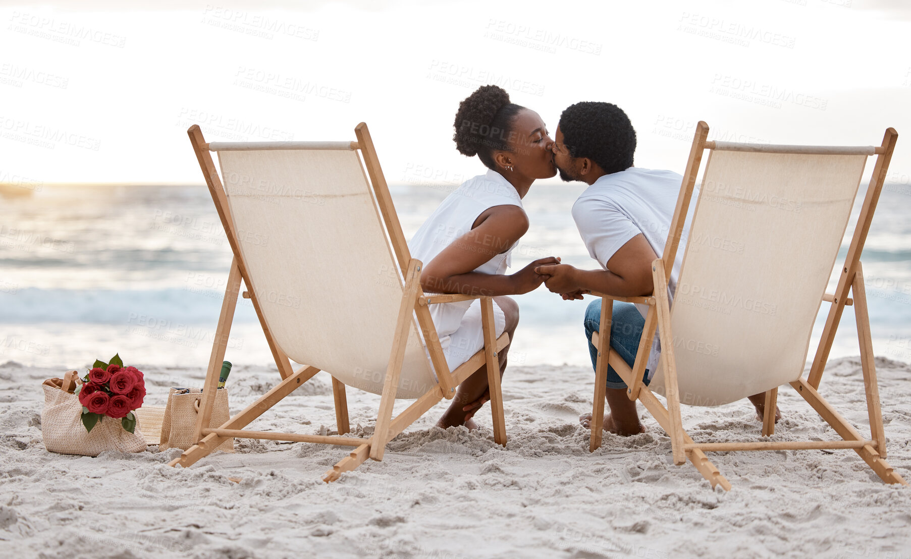 Buy stock photo Sunset, black couple and kiss with beach chair, holding hands or celebrating for anniversary. Vacation, picnic and people travel for marriage on honeymoon, summer holiday and happy bonding in Miami