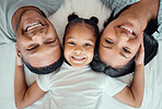 Adorable little girl pulling her parents close while lying in between her mother and father, from above. Faces of loving parents bonding with their daughter lying on a bed and spending free time together on the weekend