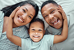 Portrait of adorable little boy with his hands on his parents paces pulling them close while lying on a bed. Cute son lying in between his mother and father, from above. Faces of loving parents bonding with their son