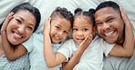 Portrait of happy mixed race family with two children relaxing and lying together on a bed at home, from above. Little brother and sister with their hands on each others faces while bonding with their loving parents