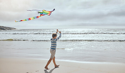 Buy stock photo Boy, child and kite at beach for vacation, playful and outdoor in wind by waves, clouds and walk by sea. Kid, toys and rainbow fabric for flight, games and holiday by ocean on adventure in Spain