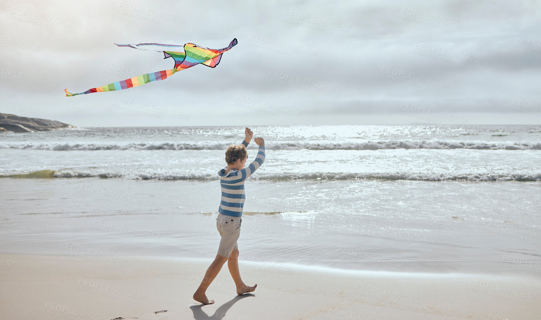 Buy stock photo Boy, child and kite at beach for vacation, playful and outdoor in wind by waves, clouds and walk by sea. Kid, toys and rainbow fabric for flight, games and holiday by ocean on adventure in Spain