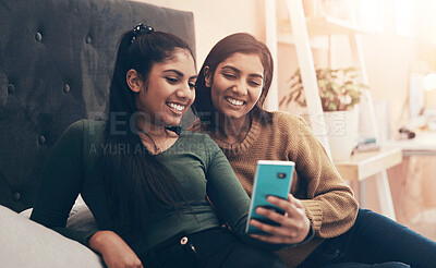 Buy stock photo Shot of two young women looking at something on a cellphone while sitting on a bed together