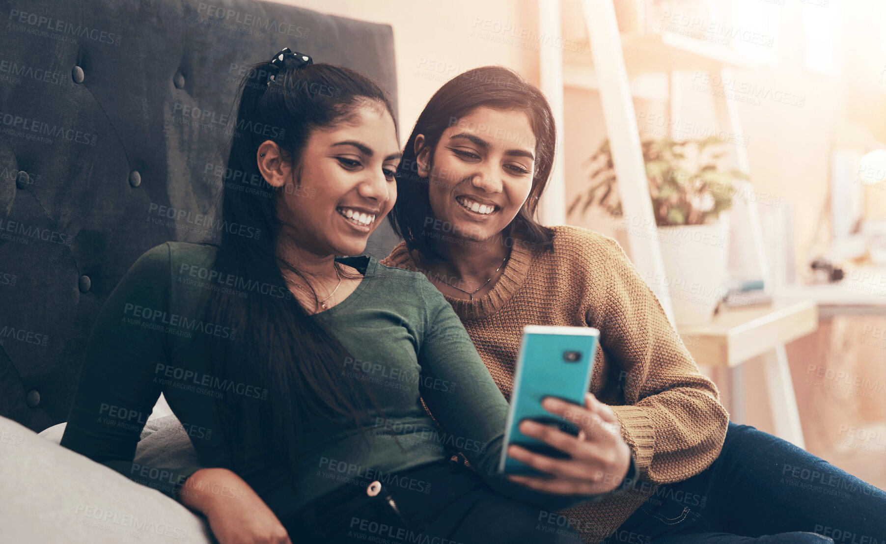 Buy stock photo Shot of two young women looking at something on a cellphone while sitting on a bed together