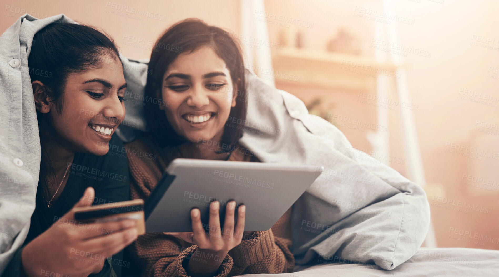 Buy stock photo Shot of two young women using a digital tablet and a credit card while lying on a bed together