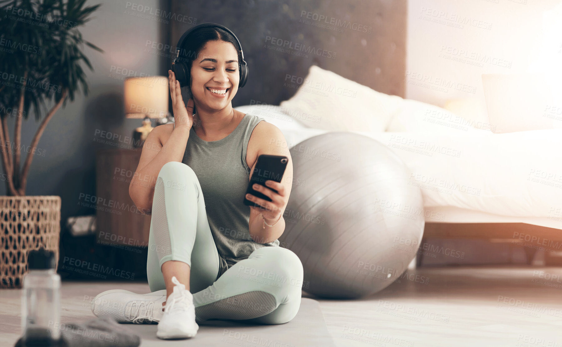 Buy stock photo Shot of a sporty young woman wearing headphones and using a cellphone while exercising at home