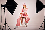 Portrait of a young mixed race female posing in trendy fashionable clothing, chilling on a chair in a grey studio background. Hispanic woman in the latest fashion collection with a cool style and hat