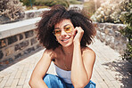 Portrait of young trendy beautiful mixed race woman with an afro smiling and posing alone outside. Hispanic woman wearing sunglasses and feeling happy. Fashionable African American woman in the city