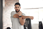 Portrait of one fit young hispanic man stretching arms and shoulders for warmup to prevent injury while exercising in a gym. Serious muscular guy focused on staying motivated and determined while mentally and physically preparing for training workout