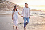 Affectionate mixed race couple holding hands and walking along the beach. Husband and wife hand in hand in the sand by the sea. Enjoying a romantic walk by the seashore at sunset. Love is in the air