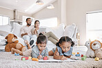 Little two girls drawing with colouring pencils lying on living room floor with their parents relaxing on couch. Little children sisters siblings colouring in during family time at home