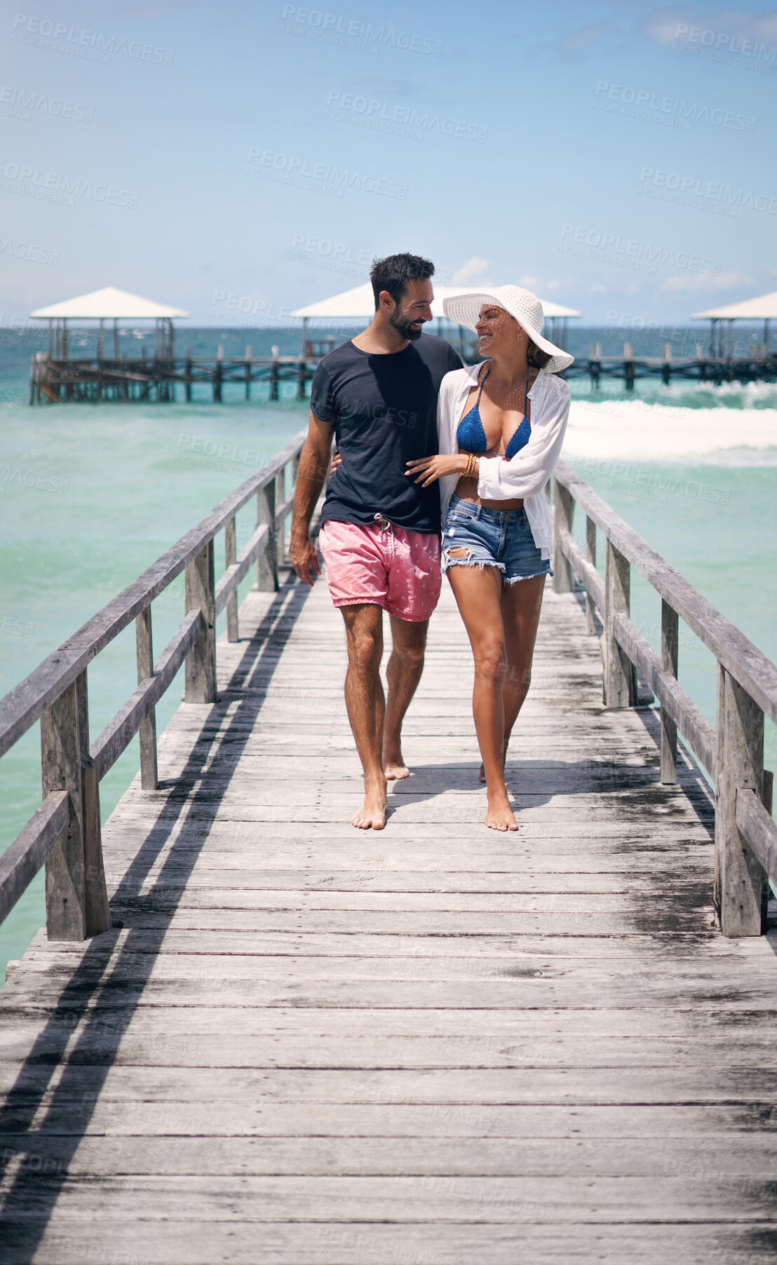 Buy stock photo Happy couple, tropical and ocean boardwalk with walk, hug and outdoor in summer sunshine for holiday. Man, woman and walking together by sea with love, happiness and talking with travel for vacation