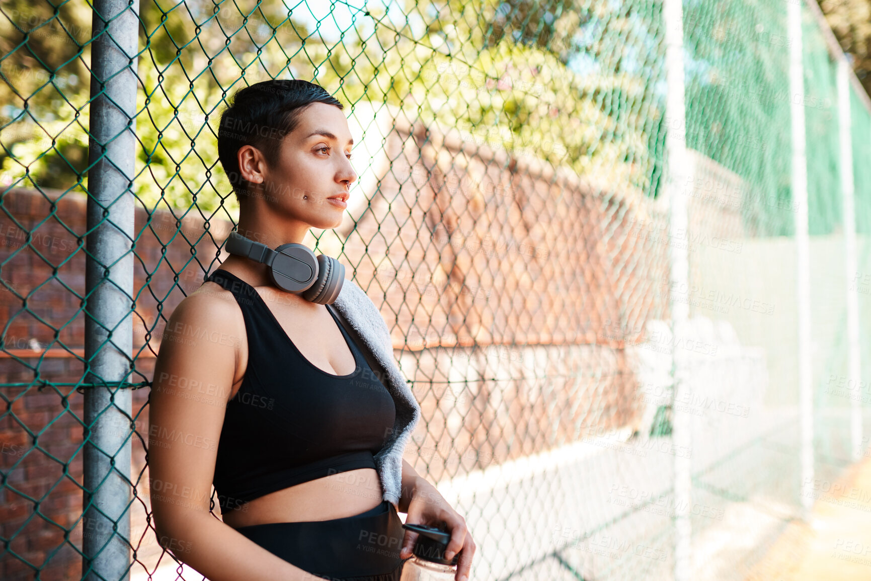 Buy stock photo Shot of a sporty young woman taking a break while standing against a fence outdoors