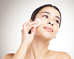 Beautiful young happy woman doing her skincare routine standing against a grey studio background alone. One content female using a cotton pad on her face while standing against a background