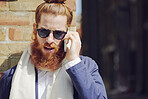 One trendy young fashionable man with man bun wearing sunglasses and talking on a cellphone while standing outside  enjoying the sun in the city. Caucasian man with a full beard against a brick wall 