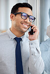 Young happy mixed race businessman on a call using a phone and  standing in an office at work. One hispanic male businessperson wearing glasses smiling while talking on the phone standing and thinking at a window in an office
