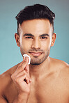 Portrait of one young indian man wiping a round cotton swab on his face while grooming against a blue studio background. Handsome mixed race guy cleaning and exfoliating his face for a healthy complexion and clear skin
