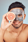 Portrait of one handsome young indian man applying a detoxifying clay or charcoal facial mask while holding grapefruit against a blue studio background. Guy using moisturising products with natural ingredients on his face for healthy, smooth soft skin