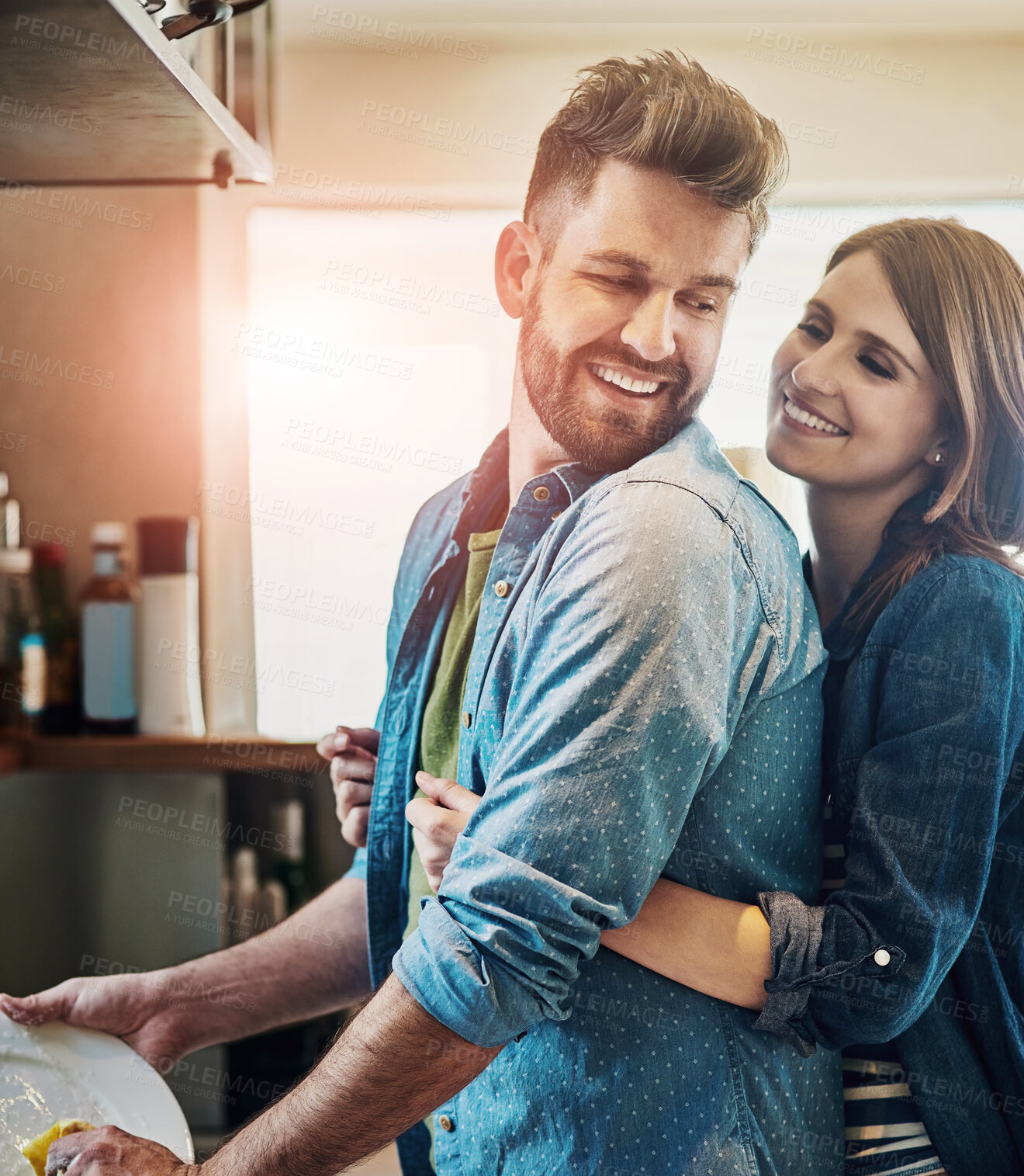 Buy stock photo Portrait of a young happy woman hugging her husband while he washes the dishes at home