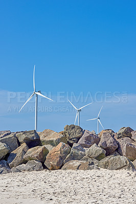 Buy stock photo Wind turbine, blue sky or beach with rocks for renewable energy, alternative power or natural sustainability in nature. Empty, fan or eco friendly environment with resource, generator or green supply