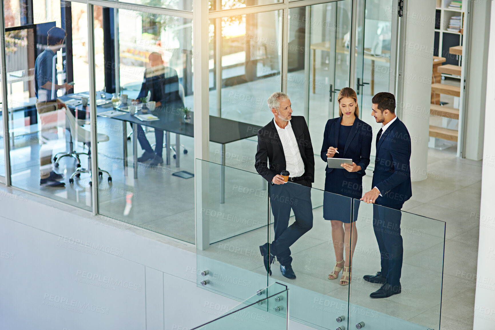 Buy stock photo Shot of three colleagues talking together over a digital tablet while standing in a modern office