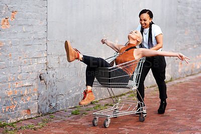 Buy stock photo Full length shot of an energetic young woman pushing her female friend in a shopping cart outdoors