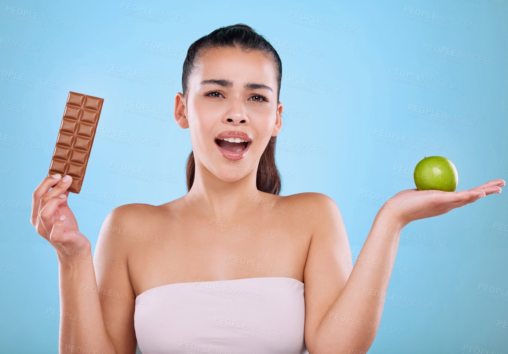 Buy stock photo Girl, chocolate and apple in studio portrait for unhealthy choice on cheat day for sweet snack for craving. Woman, isolated and blue background with fruit or cocoa, decision and balance diet.