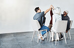 Diverse group of businesspeople giving each other a high five sitting on chairs in a meeting in an office at work. Happy women and men joining their hands celebrating success together while working