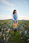 Woman farmer standing in a cabbage field on a farm. Young brunette female with a straw hat and rubber boots looking over a field of organic vegetables