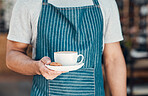 Closeup of one hispanic waiter serving a cup of coffee with a cookie while working in a cafe. Hands of barista holding teacup on saucer filled with warm latte or cappuccino for customer